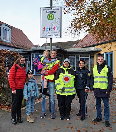 aktion_zu_fuss_zur_schule In der Ortsmitte Reisensburgs, vor dem Pfarrheim und gegenüber der Feuerwehr, ist das Schild ‚Hol- und Bringzone Grundschule‘ zu finden. Hier können die Erziehungsberechtigten ihre Kinder absetzen, diese werden dann von der Hausmeisterin oder den Schulweghelfern zur Grundschule begleitet. (V. r.: Ordnungsamtsmitarbeiter Tobias Mayer, Polizeihauptkommissarin Claudia Wecker, Hausmeisterin Waltraud Frey, Schulweghelfer Robert Eberlein mit Tochter Pauline und Schulsekretärin Silvia Schilling mit Tochter Maxima)